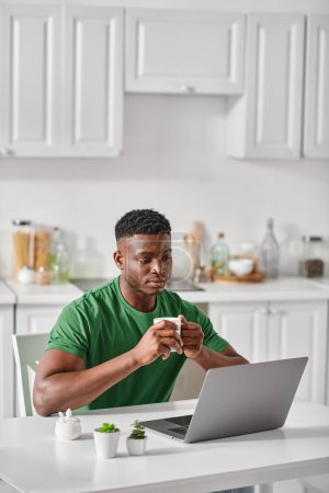 african american man enjoying cup of coffee while using laptop in kitchen, freelancer at home