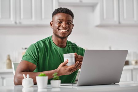 fröhlicher schwarzer Mann, der eine Tasse Kaffee genießt, während er Laptop in der Küche benutzt, Freiberufler zu Hause