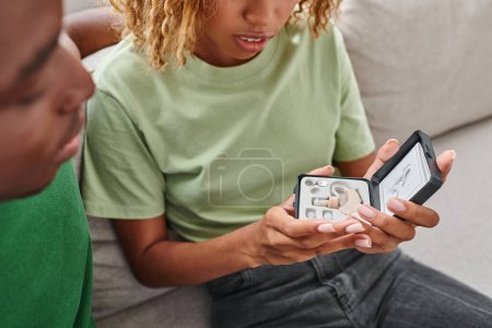 Téléchargez les photos : Femme afro-américaine cultivée tenant prothèse auditive dans une boîte près de petit ami assis sur le canapé - en image libre de droit