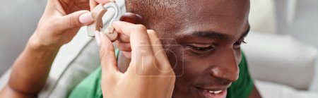 Foto de Hombre afroamericano sonriendo como su novia ayuda con audífonos, banner de equipo médico - Imagen libre de derechos