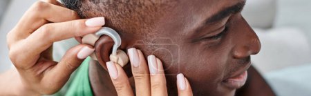 happy african american man smiling as his girlfriend assists with hearing aid, medical banner