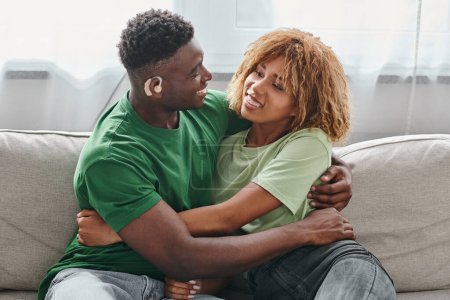 feliz pareja afroamericana abrazándose en el sofá, sordo hombre negro en audífono y mujer en aparatos ortopédicos