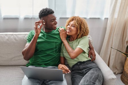hombre afroamericano feliz en dispositivo de audífono sentado en el sofá con la novia y el uso de la computadora portátil