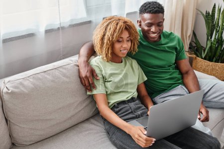 heureux homme afro-américain dans l'appareil auditif assis sur le canapé avec petite amie près d'un ordinateur portable