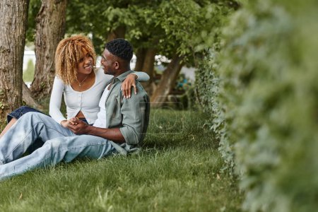 Photo for Happy african american woman in casual wear hugging boyfriend while sitting together on grass - Royalty Free Image