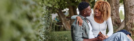 Photo for Happy african american woman in casual wear holding hands with boyfriend while sitting on grass - Royalty Free Image