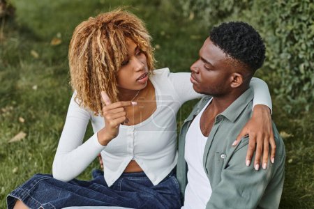 Téléchargez les photos : Jeune couple afro-américain utilisant le langage des signes pour la communication tout en étant assis sur l'herbe - en image libre de droit