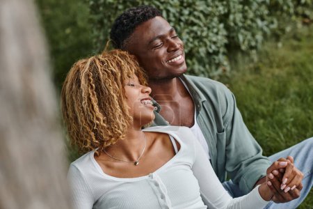 Téléchargez les photos : Excitée femme afro-américaine dans les bretelles tenant la main avec petit ami tout en étant assis dans le parc - en image libre de droit