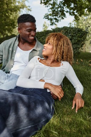 Téléchargez les photos : Joyeux couple afro-américain partageant un regard aimant tout en étant assis sur une herbe dans le parc - en image libre de droit