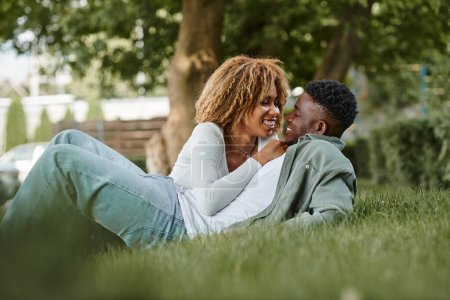 Téléchargez les photos : Joyeux jeune couple afro-américain partageant un regard affectueux tout en étant assis sur une herbe dans le parc - en image libre de droit