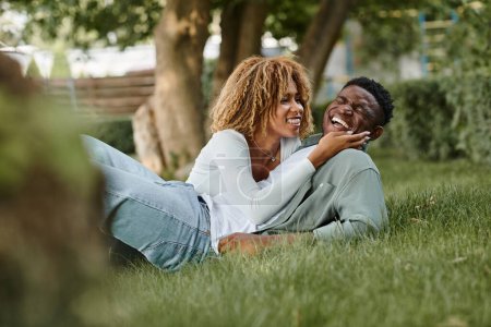 Téléchargez les photos : Joyeuse jeune femme afro-américaine partageant un regard aimant tout en étant assis sur une herbe avec l'homme - en image libre de droit