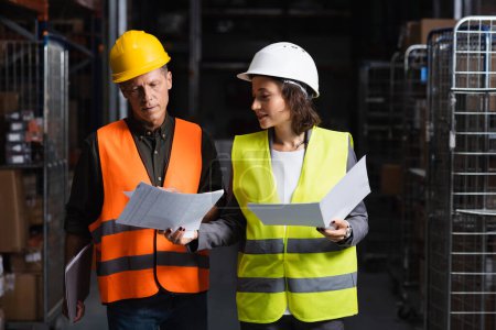 Photo for Colleagues in safety vests walking in warehouse and reviewing documents, logistic and cargo - Royalty Free Image