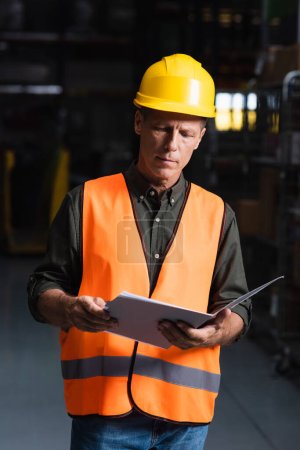Superviseur ciblé d'âge moyen en casque dur examinant la paperasserie dans l'entrepôt, la logistique et le fret