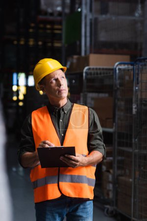 Photo for Thoughtful middle aged supervisor in hard hat reviewing paperwork in warehouse, logistics and cargo - Royalty Free Image