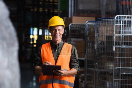 Photo for Happy middle aged supervisor in hard hat reviewing paperwork in warehouse, logistics and cargo - Royalty Free Image