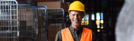 happy middle aged supervisor in hard hat reviewing paperwork in warehouse, logistics banner