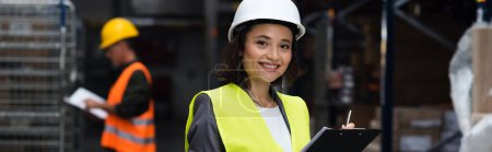 Photo for Happy warehouse worker in safety vest and hard hat writing on clipboard, logistics banner - Royalty Free Image