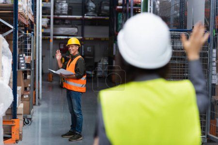 happy supervisor in hard hat waving at colleague while greeting each other in warehouse