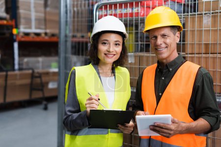 happy supervisor and employee in hard hats standing with clipboard and tablet in warehouse