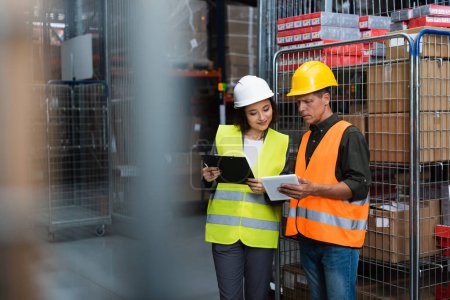 Photo for Cheerful supervisor and employee in hard hats standing with clipboard and tablet in warehouse - Royalty Free Image
