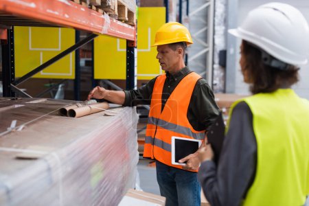 Photo for Middle aged supervisor in hard hat holding tablet while explaining work to employee in warehouse - Royalty Free Image