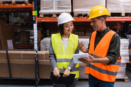 happy supervisor in hard hat holding tablet while explaining work to employee in warehouse