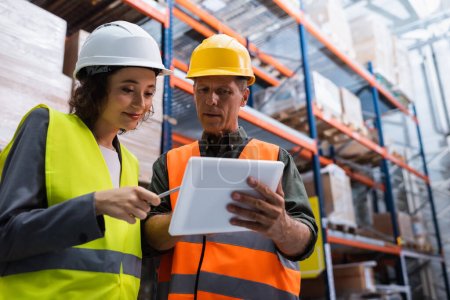 cheerful supervisor in hard hat holding tablet while explaining work to employee in warehouse