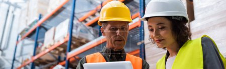 happy supervisor in hard hat holding tablet while explaining work to employee in warehouse, banner