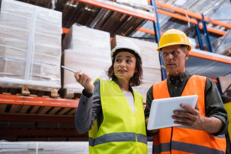 supervisor in hard hat holding tablet while discussing work to happy female employee in warehouse