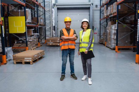 happy warehouse supervisor and employee in hard hats standing confidently and looking at camera