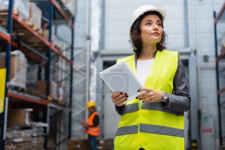 female warehouse supervisor in hard hat and safety vest holding digital tablet during work