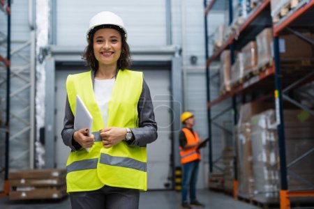 joyful female warehouse supervisor in hard hat and safety vest focused on digital tablet during work