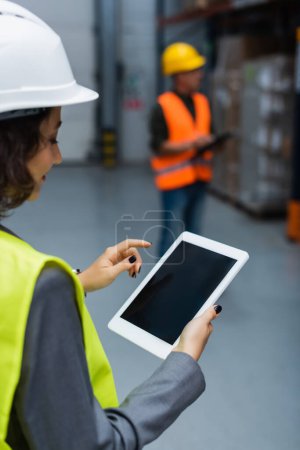 supervisora de almacén femenina en sombrero duro y chaleco de seguridad que sostiene la tableta digital con pantalla en blanco
