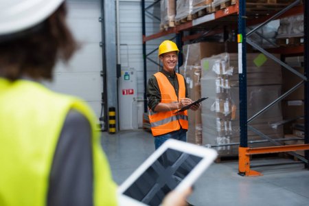 happy warehouse supervisor with clipboard and pen looking at blurred employee with tablet