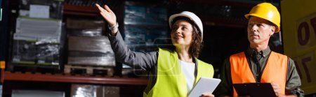 happy warehouse employee with tablet pointing away near supervisor in hard hat, horizontal banner