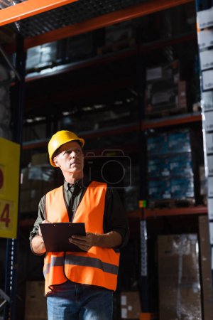 Focused middle aged warehouse supervisor in hard hat with clipboard looking at cargo on shelves