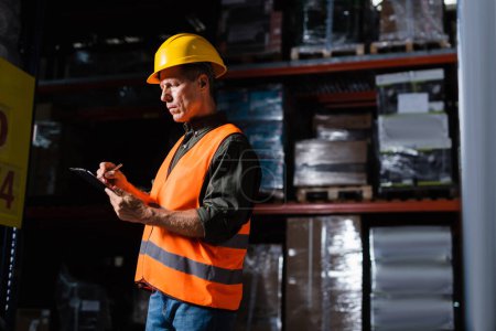 Photo for Middle aged warehouse supervisor in hard hat taking notes on clipboard while inspecting cargo - Royalty Free Image