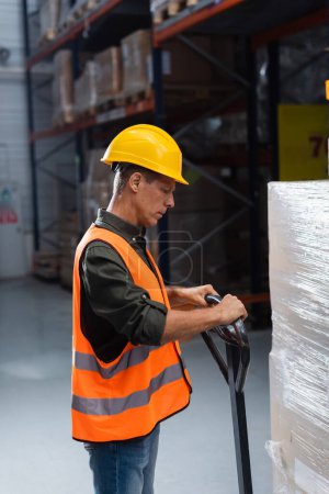 Téléchargez les photos : Travailleur d'entrepôt d'âge moyen en casque dur et gilet de sécurité transportant palette avec un camion à main - en image libre de droit