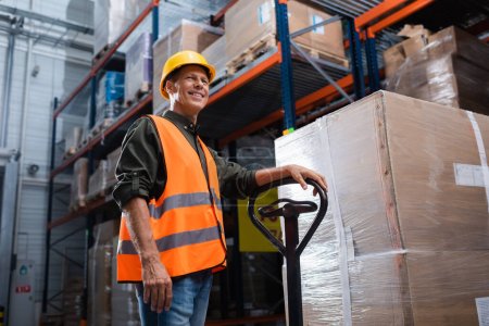 happy middle aged warehouse worker in hard hat and safety vest transporting pallet with hand truck