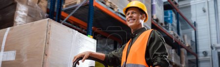happy middle aged warehouse worker in safety vest transporting pallet with hand truck, banner