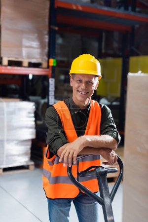 travailleur d'entrepôt joyeux en casque dur et gilet de sécurité transportant palette avec camion à main, sourire