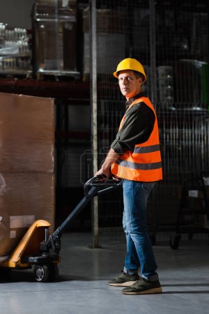 strong middle aged warehouse worker in safety vest transporting heavy pallet with hand truck