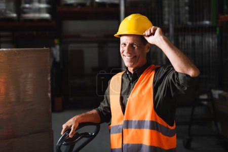 cheerful middle aged warehouse worker in safety vest smiling near hand truck, professional headshot