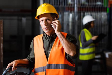 Photo for Warehouse supervisor making a phone call on smartphone with a female colleague in the background - Royalty Free Image