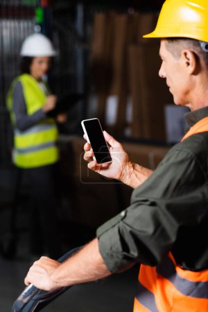 Foto de Supervisor en chaleco de seguridad y casco usando smartphone con empleado en segundo plano del almacén - Imagen libre de derechos