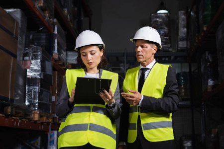 middle aged supervisor discussing work with female employee writing on clipboard in warehouse