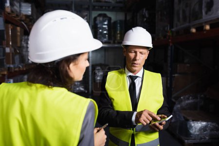 Photo for Middle aged supervisor in hard hat using smartphone near female employee on blurred background - Royalty Free Image