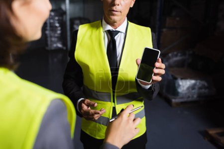 Photo for Cropped middle aged supervisor in safety vest using smartphone near blurred female employee - Royalty Free Image