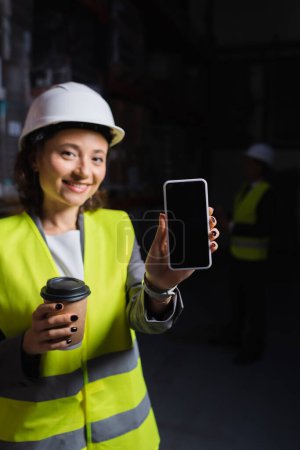 smiling woman in hard hat holding smartphone and paper cup, happy warehouse employee on coffee break