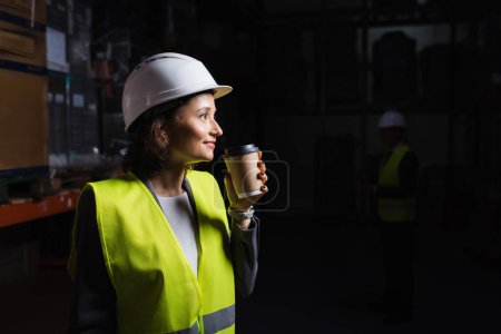 Téléchargez les photos : Vue latérale de la femme en chapeau dur tenant tasse en papier, employé heureux entrepôt pendant la pause café - en image libre de droit